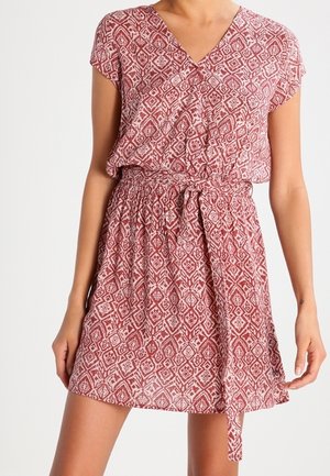 Woman wearing a red patterned short-sleeve wrap dress with a waist tie, standing with arms relaxed against a plain white background.