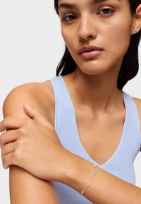 Pulsera de plata con pequeñas piedras preciosas transparentes, llevada en un brazo bronceado. La modelo lleva una camiseta sin mangas de canalé en azul claro, mostrando la pulsera.