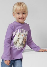 Jong meisje met blond haar dat glimlacht, gekleed in een paarse sweatshirt met drie paarden en de tekst "MEISJES VRIENDEN wie regeert de wereld? MEISJES!"