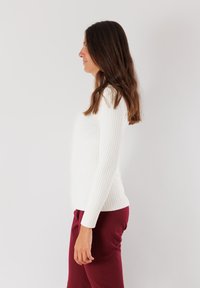 Top a maniche lunghe a coste bianche con una silhouette aderente, abbinato a pantaloni bordeaux. Tessuto liscio e scollatura regolare. Vista laterale.
