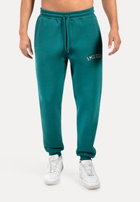 Tyrkysové tepláky s elastickým pasem a šňůrkou, s bočními kapsami a vyšitým logem "SMILDOODX" na levém stehně.