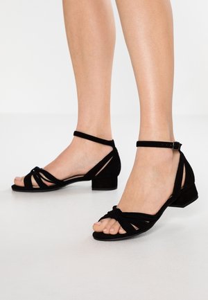 Jambes portant des sandales noires à petits talons avec des brides à la cheville et des brides avant nouées sur un sol et un fond blancs.