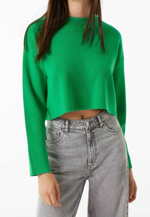Femme portant un pull court vert vif et un jean taille haute gris clair, debout avec une main détendue le long du corps.