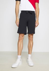 Pantaloni corti neri in cotone con elastico in vita e stringa, dotati di tasche laterali con zip e taglio dritto. Abbinati a sneakers bianche.