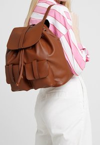 Brauner Leder-Rucksack mit Schnürzug und Klappenverschluss, der zwei Außentaschen und eine strukturierte Oberfläche aufweist.