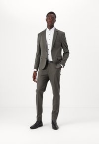 Costume gris au design ajusté, veste à un bouton et pantalon assorti. Comprend une chemise blanche avec des boutons noirs, portée avec des chaussures noires.