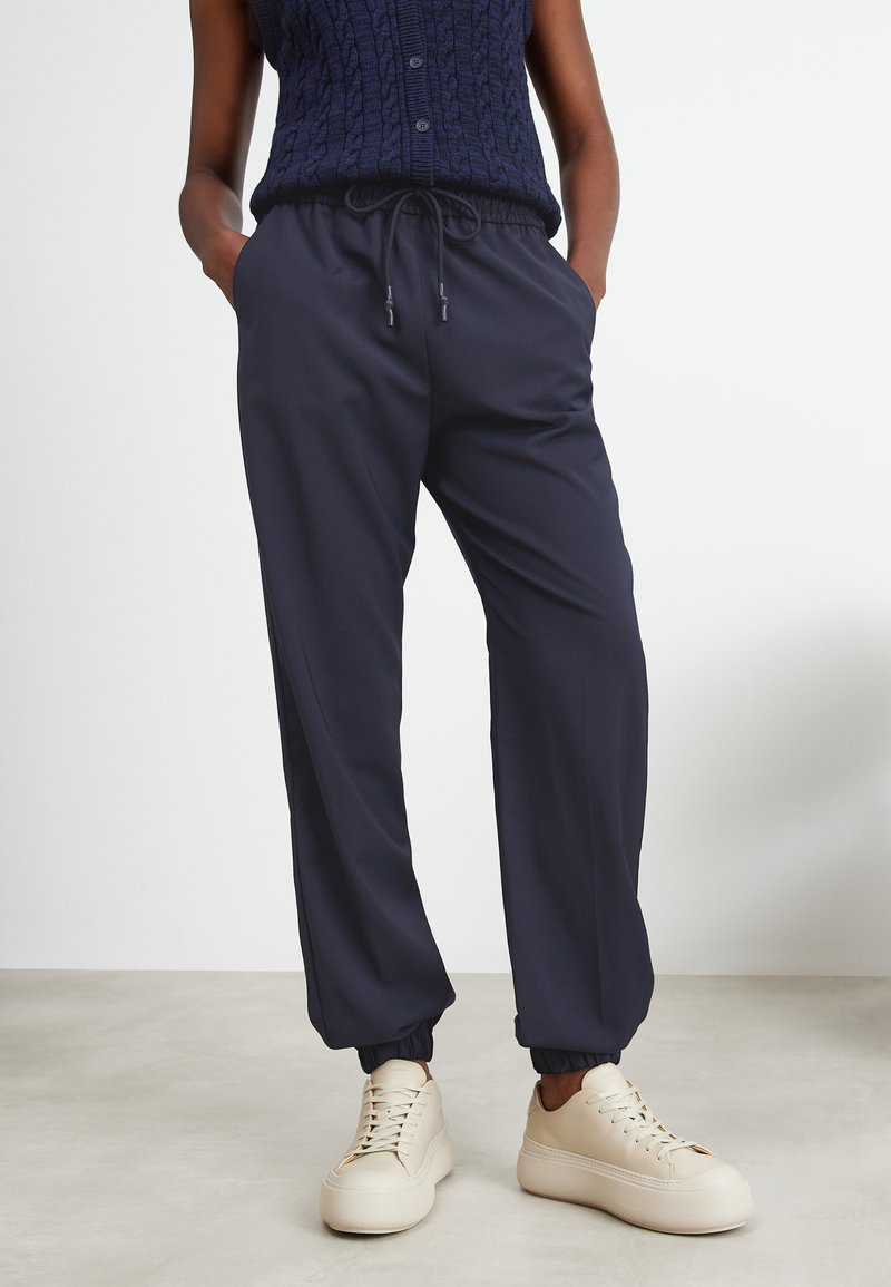 Osoba w granatowych spodniach dresowych z gumką i kremowych trampkach na platformie, stojąca z rękami w kieszeniach na jasnej podłodze.