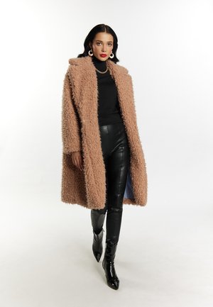 Cappotto oversize, beige, in pelliccia sintetica con una finitura testurizzata; indossato su un dolcevita nero e abbinato a pantaloni in pelle neri e stivaletti a punta.