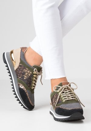 Personne portant un pantalon blanc et des baskets à textures multiples avec des accents verts, noirs, beiges, dorés, et un tissu à motifs, lacées et debout sur un sol gris.