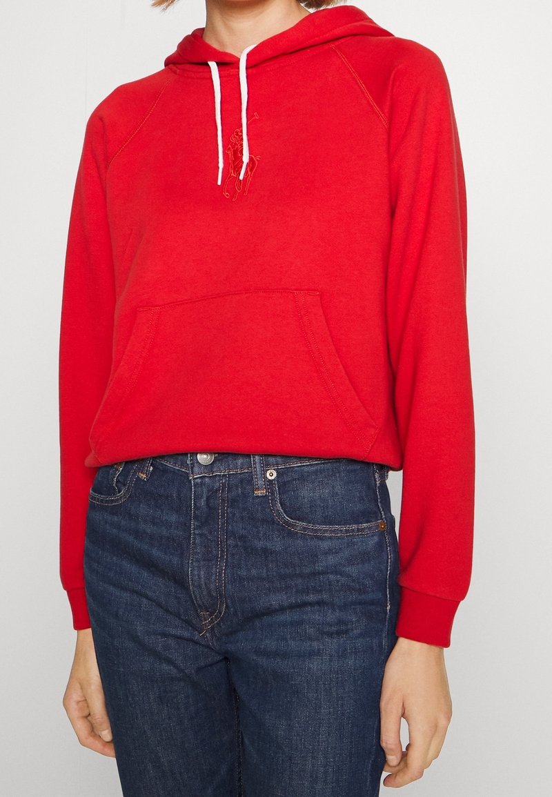 Personne portant un sweat à capuche rouge avec des cordons blancs et une poche à l'avant, assorti à un jean bleu foncé, visible des épaules aux hanches.