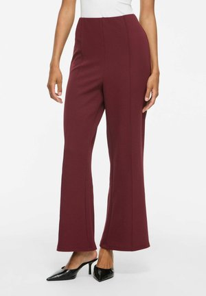 Partie inférieure du corps d'une personne portant un pantalon large taille haute bordeaux avec des mules à talons noires à bout pointu sur un fond blanc uni.