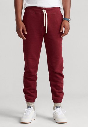 Personne portant un pantalon de jogging bordeaux avec un cordon blanc, des chaussettes beige, des baskets blanches, une chemise blanche et une montre bleue au poignet gauche.