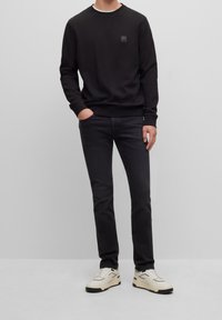 Hombre vestido con sudadera negra y jeans negros, con zapatillas blancas, de pie frente a un fondo gris claro liso.