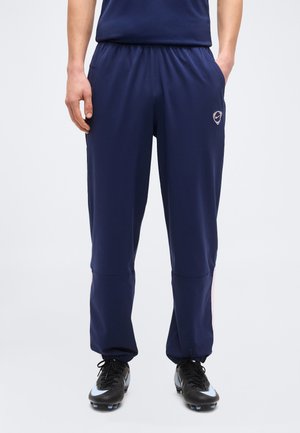 Mann iført marineblå treningsbukser med elastiske mansjetter og svarte fotballsko med lyseblå detaljer, stående med hendene i lommene.