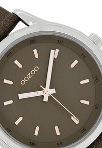 Brown leather strap watch with a silver stainless steel case. Dark brown dial features gold and white hour markers and hands. Brand name "OOZOO."