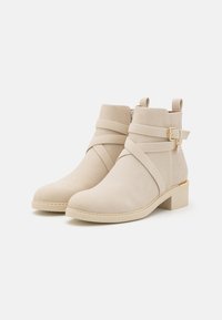 Bottines beiges en daim doux avec des brides croisées et une petite boucle latérale. Dotées d'un talon bloc et d'un panneau arrière texturé.