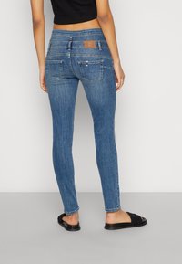 Mujer vistiendo jeans ajustados azules, top corto negro y sandalias negras de tiras, de pie contra un fondo gris claro liso.