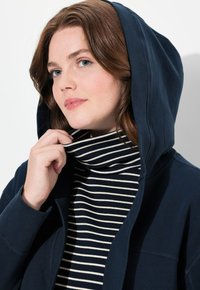 Marinblå hoodie med mjuk textur och fodrad huva, bärs över en randig polotröja. Polotröjan har vita och marinblå ränder.