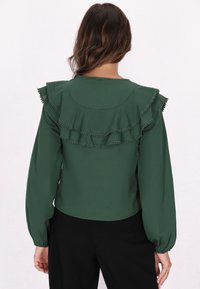 Grüne Bluse mit runden Ausschnitt, langen Puffärmeln und geschichteten Rüschen entlang des Rückens, akzentuiert mit kleinen Besatzdetails.
