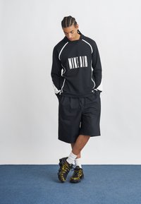 Melnā sporta krekls ar "NIKE AIR" zīmolu, baltām akcentiem, apvienots ar melnām šortiem un melni-dzelteniem sporta apaviem. Gluda auduma, brīvs piegriezums.