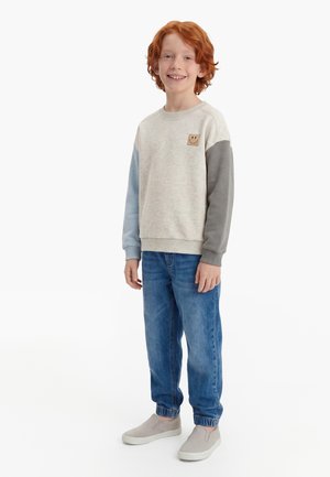 Rothaariges Kind lächelt, trägt einen beigen Pullover mit einem Smiley-Patch, blaue Jeans und beige Slip-On-Schuhe, steht vor weißem Hintergrund.
