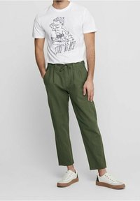 Witte graphic t-shirt met een cartoonpersonage, gecombineerd met groene broek met aantrekkoord en witte sneakers met bruine accenten.