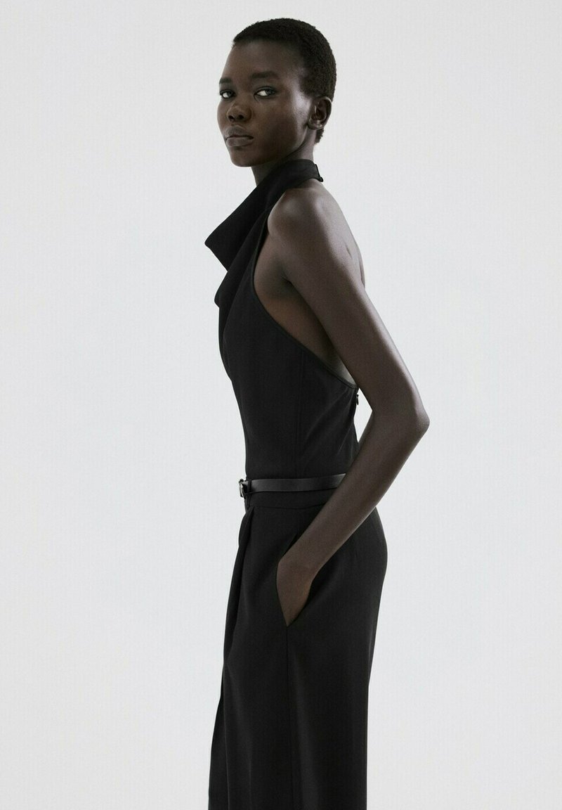 Femme aux cheveux courts portant une combinaison noire sans manches, posant de profil avec une main dans la poche sur un fond blanc uni.