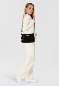 Borsa a tracolla nera con hardware color oro, texture liscia, caratterizzata da un accento dorato e un charm in pelle laterale, mostrata su uno sfondo chiaro.