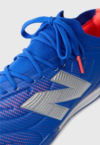 Sapatilha desportiva azul com parte superior texturizada, logótipo "N" prateado e detalhes em vermelho. Apresenta uma biqueira arredondada e atacadores trançados.