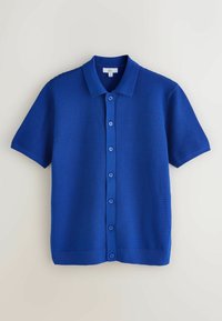 Chemise en maille texturée bleue à manches courtes, boutonnée et avec col, étalée à plat sur une surface claire.