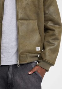 Giacca bomber verde oliva con finitura strutturata, chiusura con zip, polsini a coste e tasche frontali. Include una piccola etichetta con logo sul lato.