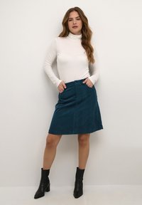 Teal corduroy skirt with side pockets, button closure, and a knee-length hem, paired with a fitted white turtleneck and black ankle boots.