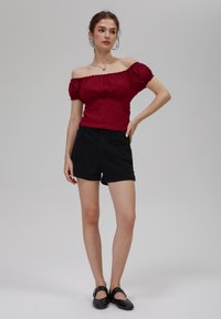 Rød off-shoulder topp med teksturert mønster, kombinert med svarte denimshorts. Har korte ermer og en tettsittende design. Svarte flate sko.