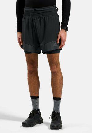 Atleet draagt zwarte dubbele hardloopshorts, grijze enkelsokken, zwarte hardloopschoenen en een zwart shirt met lange mouwen, rechtopstaand.