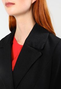 Svart textured blazer med snedkrage, bärs över en röd topp, framhäver en strukturerad design och skarpa linjer.