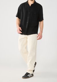 Maglietta nera a maniche corte con chiusura a bottoni e taschino, abbinata a pantaloni larghi color crema e sneakers nere con strisce bianche.