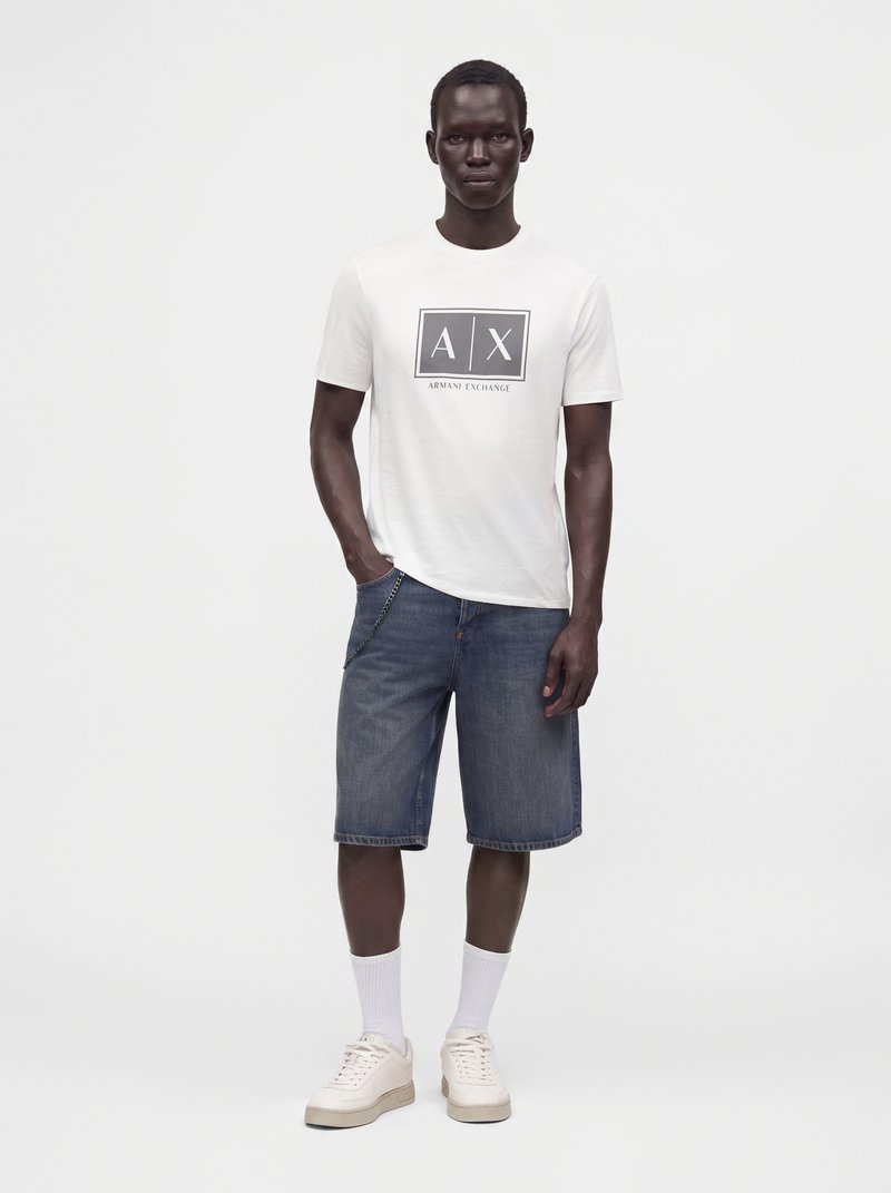 Мъж стои, облечен с бяла тениска с лого на Armani Exchange, сини дънкови шорти, бели чорапи и бежови маратонки пред едноцветен бял фон.