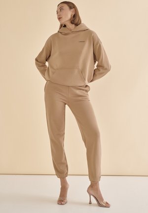 Beige kapucnis pulóver, elülső zsebből és hozzá illő joggersből áll. Puha anyagú textúra, szűkített mandzsetták, amelyeken egy kis logó látható. Tiszta sarkú cipők viselése.