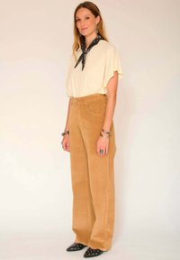T-shirt beige oversize, pantalon en velours côtelé marron à jambes larges, et chaussures noires à bout pointu avec des accents en argent. Écharpe nouée au cou.