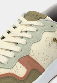 Αθλητικό παπούτσι σε ανοιχτό χρώμα με υφασμάτινο κρεμ επάνω μέρος, πράσινες και σκουρόχρωμες καφέ λεπτομέρειες και κρεμ κορδόνια. Το χρυσό λογότυπο της μάρκας είναι ορατό στο πλάι.