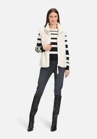 Gilet in pile crema con colletto alto sopra una maglietta a righe bianche e nere a maniche lunghe. Il modello indossa jeans scuri e stivali neri sopra il ginocchio.