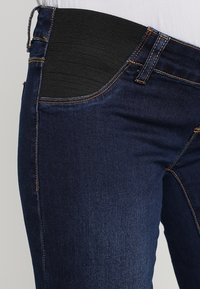 Vaqueros de maternidad de color azul oscuro con un panel elástico en la cintura y detalles de costura en naranja en el bolsillo y las costuras.