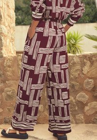Pantalon à jambes larges couleur marron avec un motif géométrique à rayures blanches et une taille élastique. Tissu texturé avec une coupe décontractée.