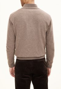 Maglione lavorato a maglia marrone con colletto alto, con polsini e orlo a coste. Indossato con pantaloni in velluto a coste marroni scuri, mostrato da dietro.