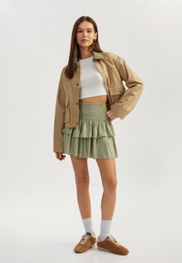 Veste beige raccourcie, haut blanc ajusté, jupe verte à volants et baskets marron. Les accessoires comprennent des chaussettes blanches et des boucles d'oreilles discrètes.
