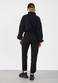 Μαύρη, υπερμεγέθη φόρμα με ψηλό γιακά και ελαστικά τελειώματα, συνδυασμένη με μαύρα αθλητικά παπούτσια. Μινιμαλιστικός σχεδιασμός, λείο υλικό.