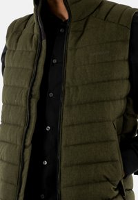 Gilet verde imbottito con chiusura a zip, texture trapuntata e tasche laterali, indossato sopra una camicia nera con bottoni. Logo impresso sul tessuto.