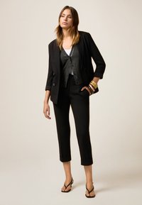 Blazer nero su misura con maniche ripiegate sopra un gilet scuro, abbinato a pantaloni cropped neri slim e sandali neri con cinturini. Braccialetti d'oro in evidenza.