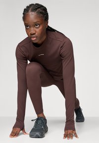 Giovane donna con capelli intrecciati, vestita con abbigliamento sportivo marrone e scarpe da corsa nere, in posizione di partenza per uno sprint su uno sfondo semplice.