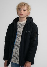 Svart, huva pufferjacka med dragkedja och sidofickor, lagrad över en krämfärgad skjorta med svarta abstrakta textmönster.